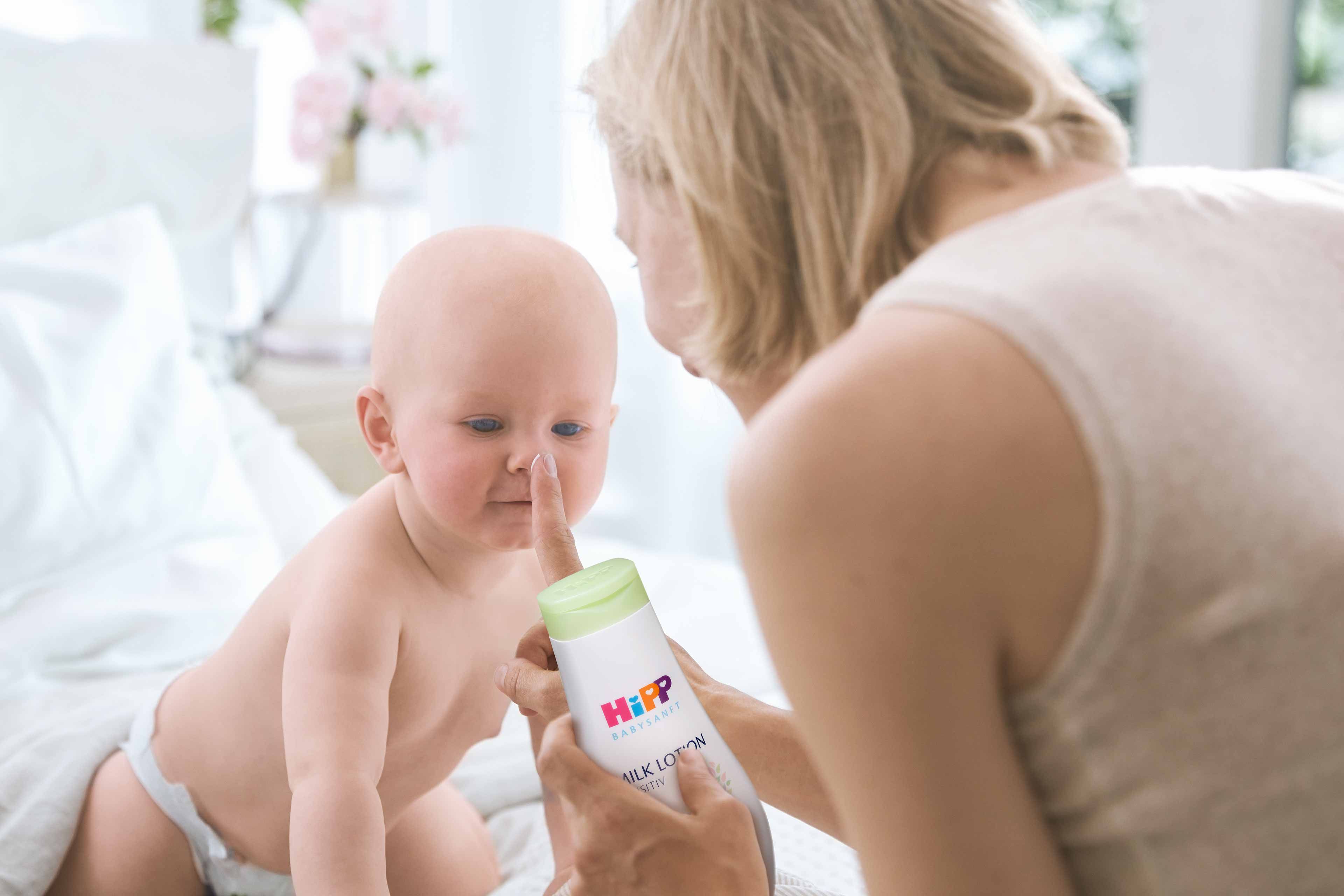 Ein Baby, das neugierig zu sein scheint, spielt mit einer Flasche mit der Aufschrift "Hipp Milk Lotion", die von einer Frau gehalten wird.