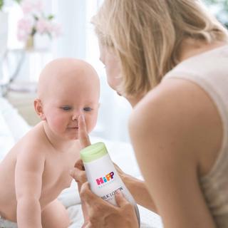 Ein Baby, das neugierig zu sein scheint, spielt mit einer Flasche mit der Aufschrift "Hipp Milk Lotion", die von einer Frau gehalten wird.