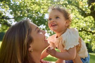 Lachende Mutter hält ihr Kleinkind im grünen Park im Arm, beide schauen sich liebevoll an und genießen die gemeinsame Zeit.
