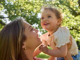 Lächelnde Mutter hält ihr fröhliches Kleinkind im Grünen – liebevoller Moment wie im HiPP Mein BabyClub