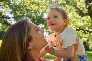 Lachende Mutter hält ihr Kleinkind im grünen Park im Arm, beide schauen sich liebevoll an und genießen die gemeinsame Zeit.