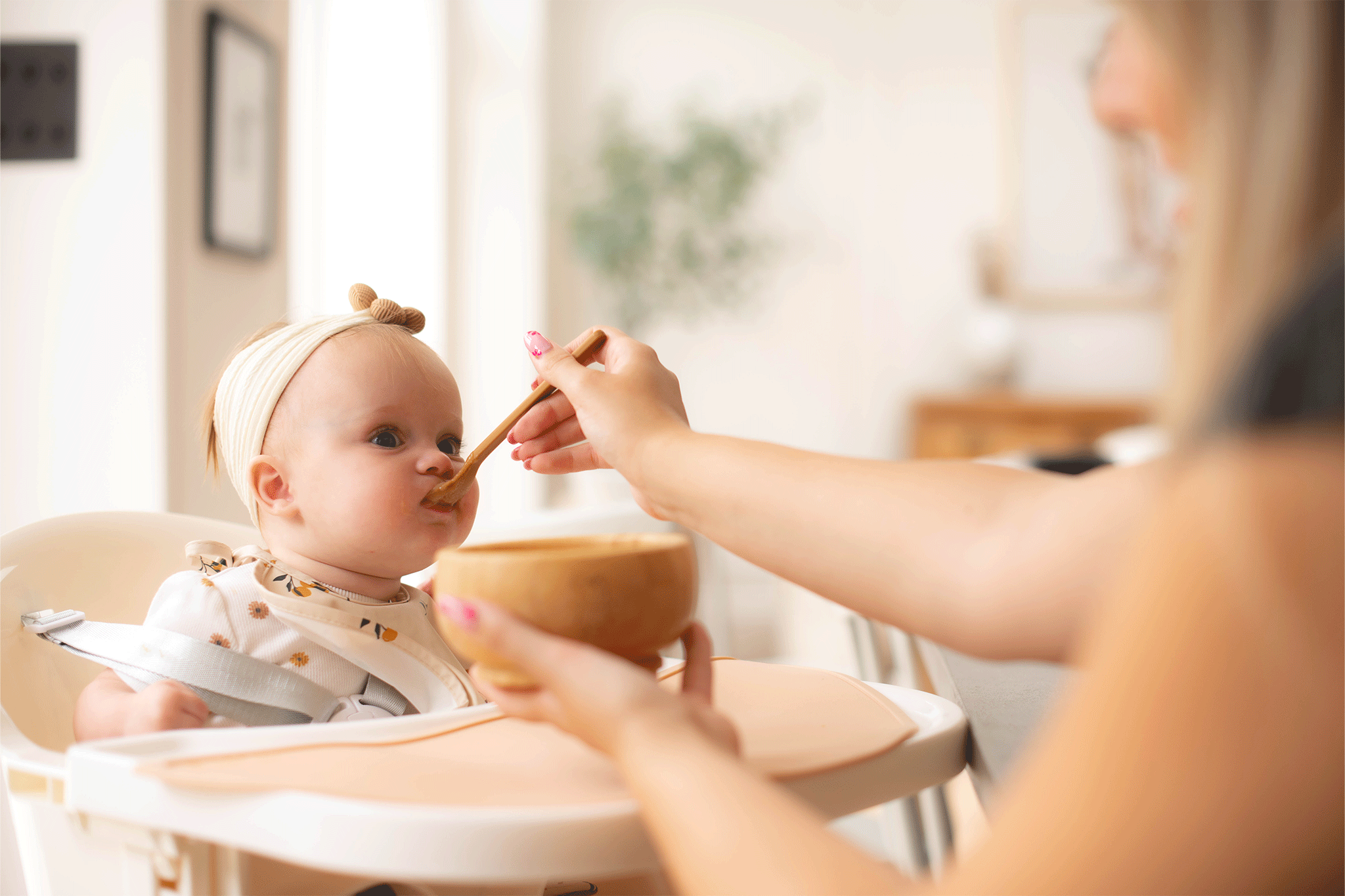 A baby sitting in a high chair wearing a knot headband and bib while an adult off-camera feeds them food from a wooden bowl with a wooden spoon in a brightly lit room.