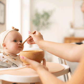 A baby sitting in a high chair wearing a knot headband and bib while an adult off-camera feeds them food from a wooden bowl with a wooden spoon in a brightly lit room.