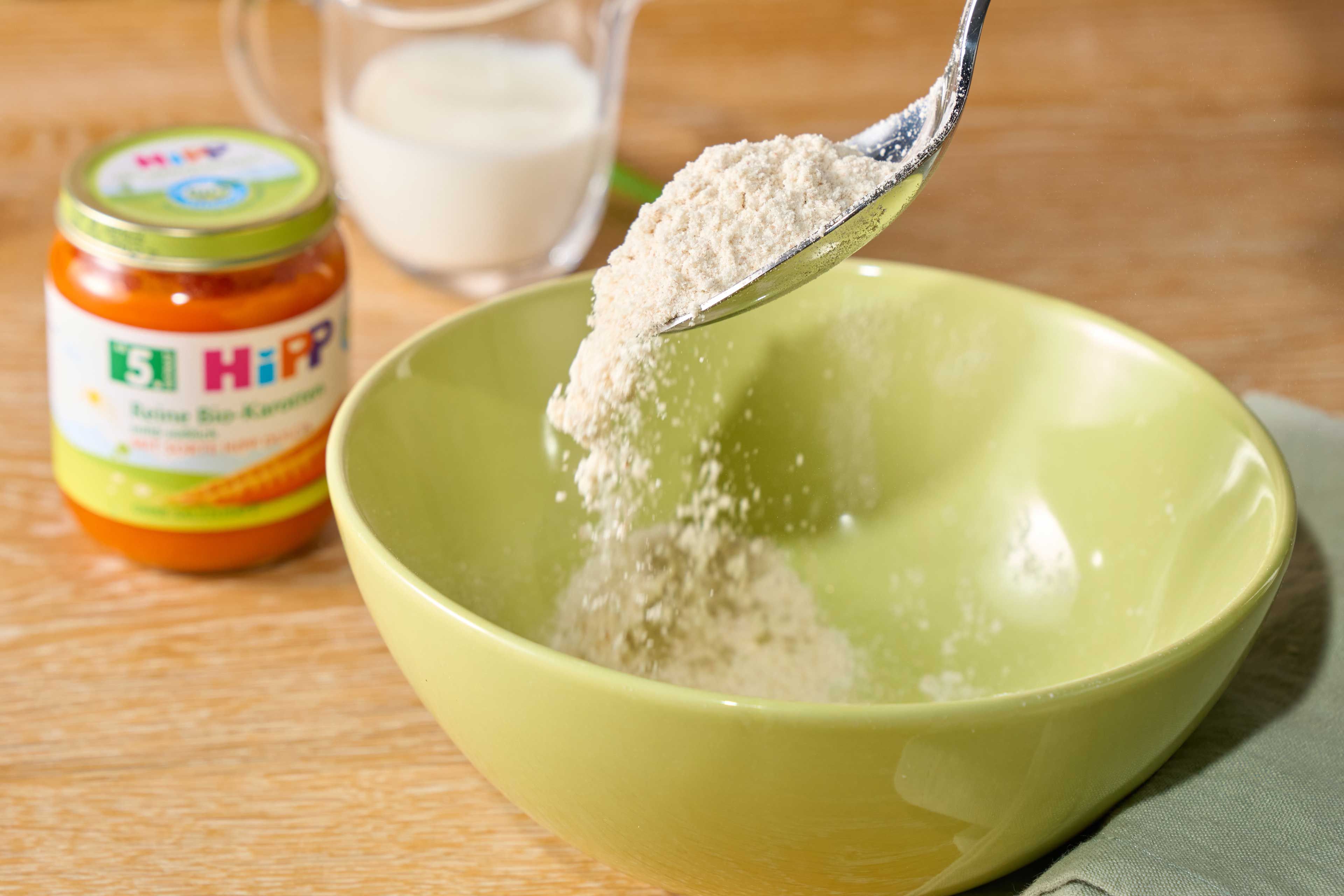 Löffel schüttet Mehl in eine grüne Schüssel, im Hintergrund steht ein Glas mit weißem Pulver und ein Glas mit Babybrei der Marke HiPP