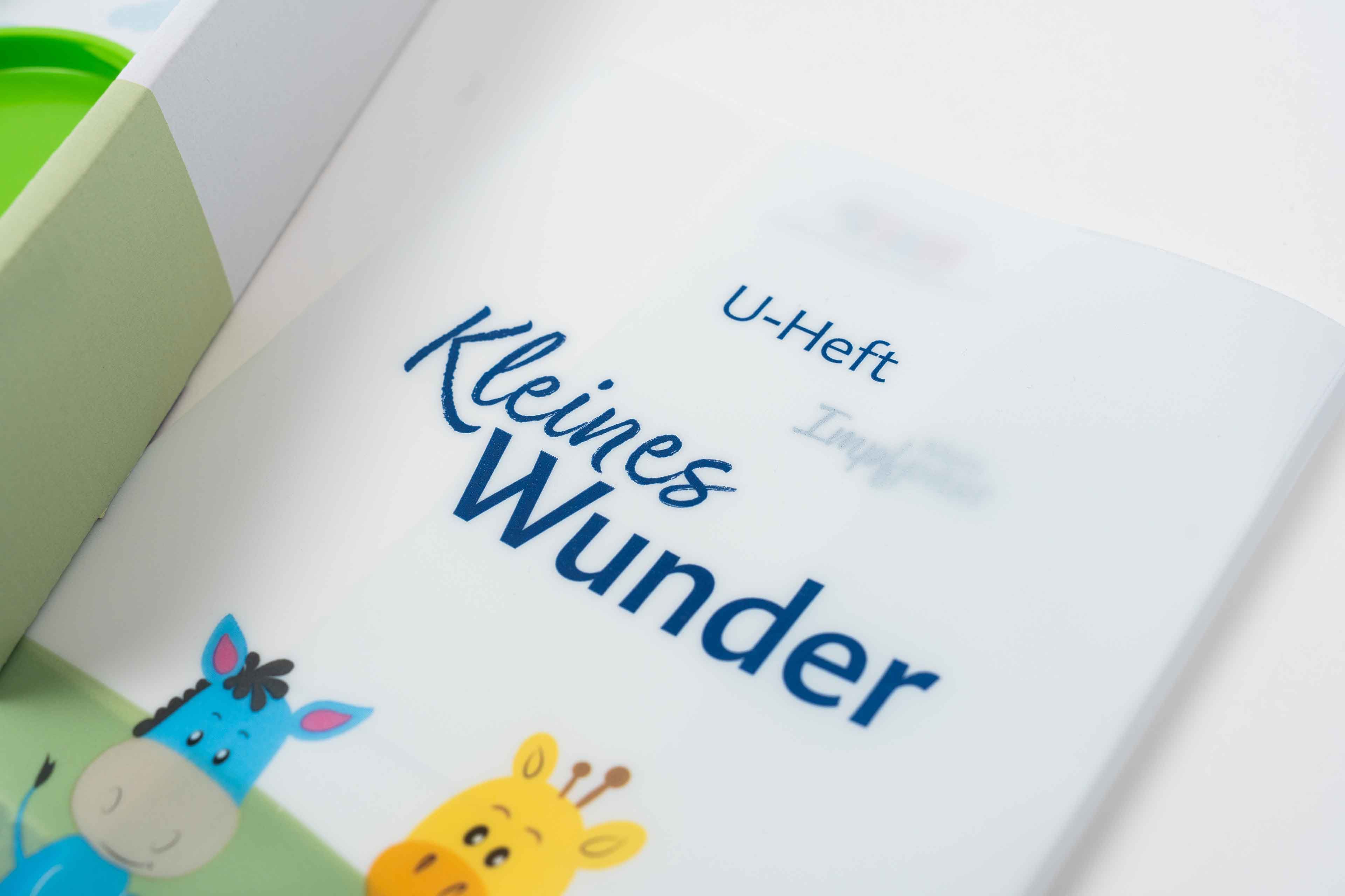 Weiße U-Heft-Hülle mit der blauen Aufschrift 'U-Heft Kleines Wunder' und Illustrationen eines blauen Esels und einer gelben Giraffe am unteren Rand