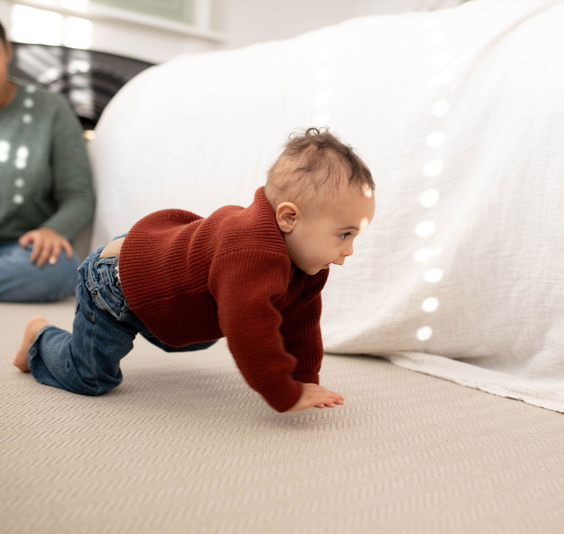 Baby in a red jumper crawling on a soft carpet while a parent watches nearby in a bright, cosy room.