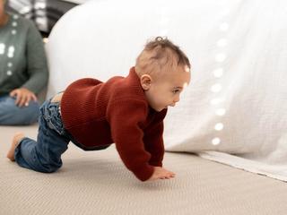 Baby in a red jumper crawling on a soft carpet while a parent watches nearby in a bright, cosy room.