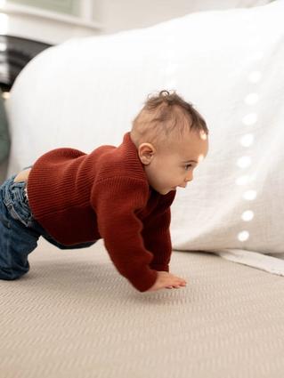 Baby in a red jumper crawling on a soft carpet while a parent watches nearby in a bright, cosy room.