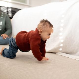 Baby in a red jumper crawling on a soft carpet while a parent watches nearby in a bright, cosy room.