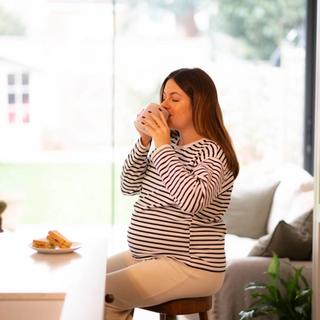 A pregnant woman in a striped top sits on a stool, sipping from a mug in a bright kitchen. A plate with sandwiches rests on the counter beside her, and soft natural light fills the cozy space.