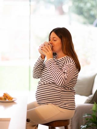 A pregnant woman in a striped top sits on a stool, sipping from a mug in a bright kitchen. A plate with sandwiches rests on the counter beside her, and soft natural light fills the cozy space.