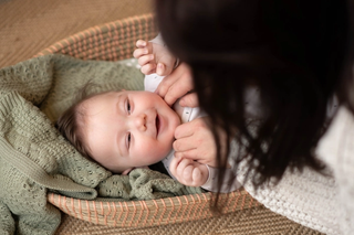 A smiling baby lies on a green knitted blanket inside a woven basket, looking up joyfully while an adult gently tickles their neck. The cozy scene captures a sweet bonding moment on a textured rug