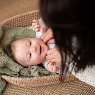 A smiling baby lies on a green knitted blanket inside a woven basket, looking up joyfully while an adult gently tickles their neck. The cozy scene captures a sweet bonding moment on a textured rug.