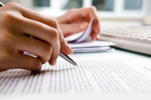 Die Hände einer Person, eine hält einen Stift, die andere einige Papiere, mit einem unscharfen Hintergrund einer Tastatur und eines Dokuments.