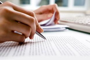 Die Hände einer Person, eine hält einen Stift, die andere einige Papiere, mit einem unscharfen Hintergrund einer Tastatur und eines Dokuments.