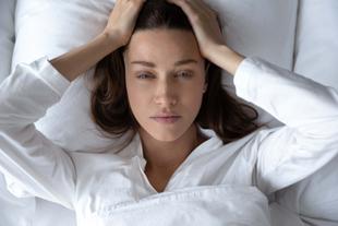 Eine Frau in Weiß liegt auf einem Bett, die Hände auf dem Kopf.