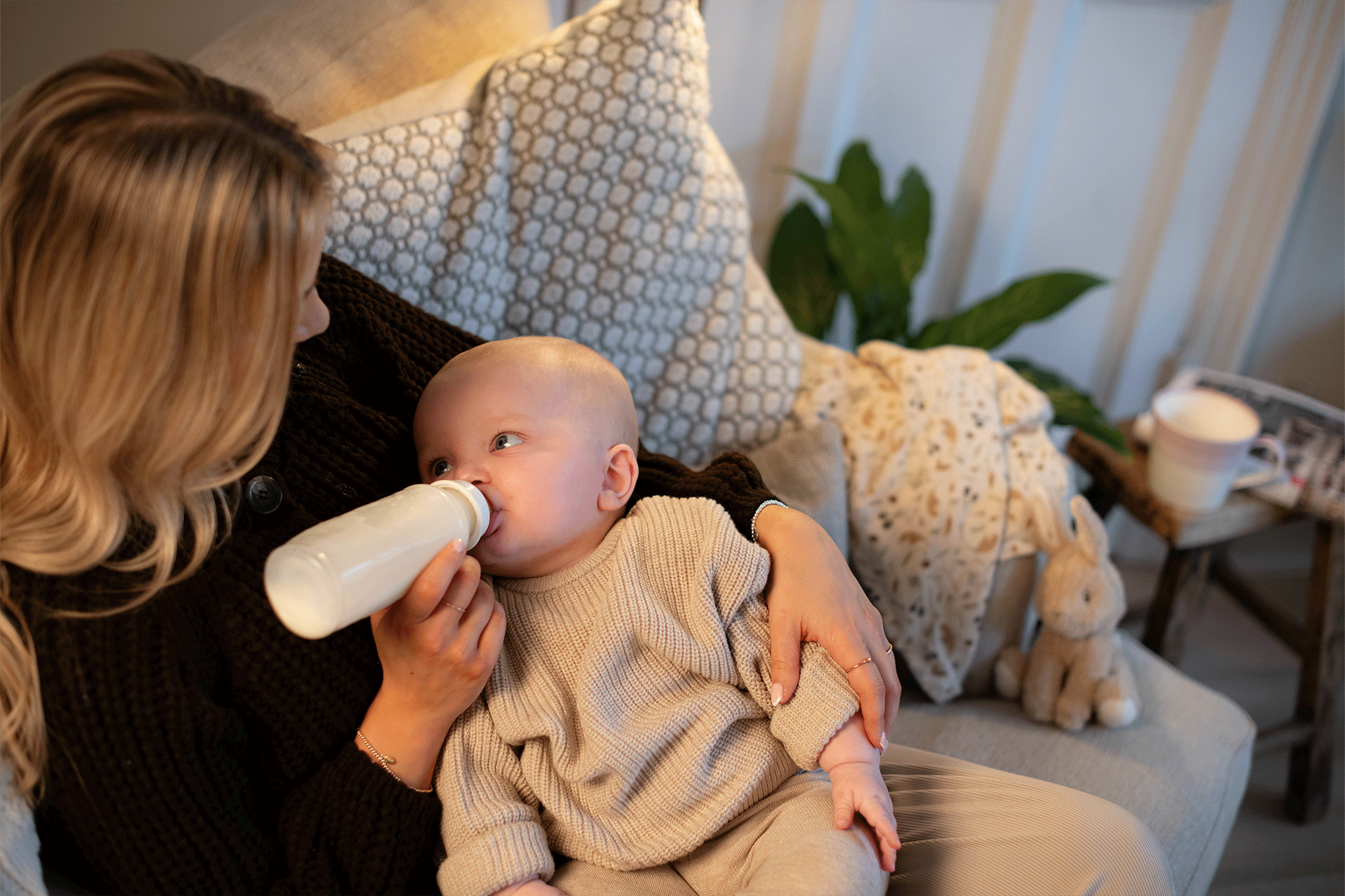 A blonde woman wearing a dark knit cardigan sits on a light gray sofa and bottle-feeds her baby. The baby, dressed in a ribbed beige sweater and pants, lies cradled in her arms looking up at her while drinking from a clear baby bottle filled with milk. Beside them on the sofa is a stuffed bunny toy, patterned pillows, and a blanket, with a mug on a small stool in the background.