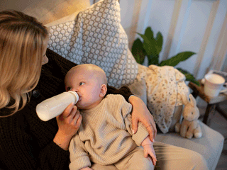 A blonde woman wearing a dark knit cardigan sits on a light gray sofa and bottle-feeds her baby. The baby, dressed in a ribbed beige sweater and pants, lies cradled in her arms looking up at her while drinking from a clear baby bottle filled with milk. Beside them on the sofa is a stuffed bunny toy, patterned pillows, and a blanket, with a mug on a small stool in the background.