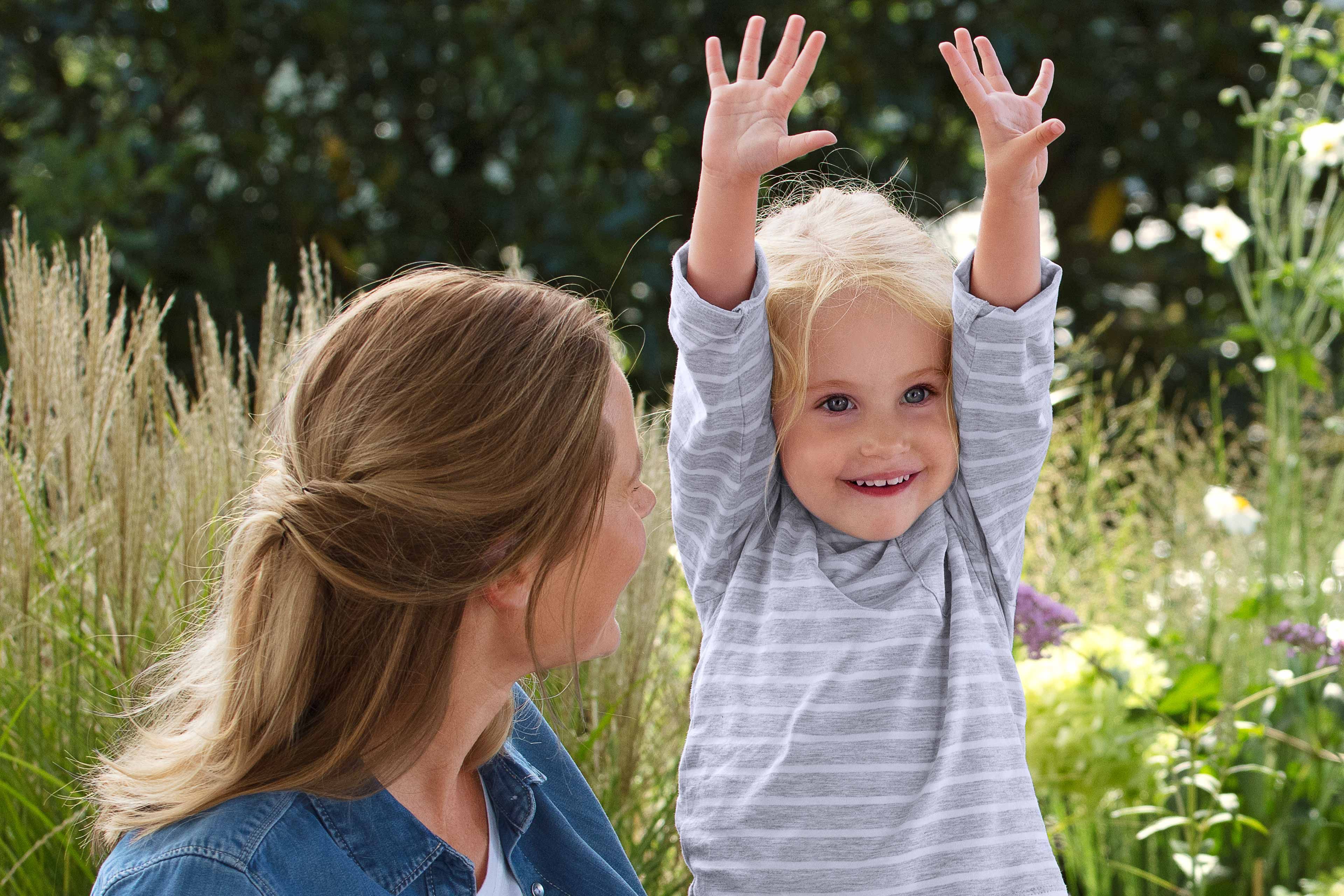 Ein junges Mädchen mit blondem Haar, das in einer spielerischen Geste die Hände hebt, umgeben von einem Garten mit weißen Blumen und Grünzeug.