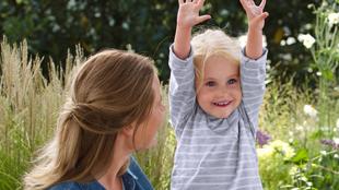 Ein junges Mädchen mit blondem Haar, das in einer spielerischen Geste die Hände hebt, umgeben von einem Garten mit weißen Blumen und Grünzeug.