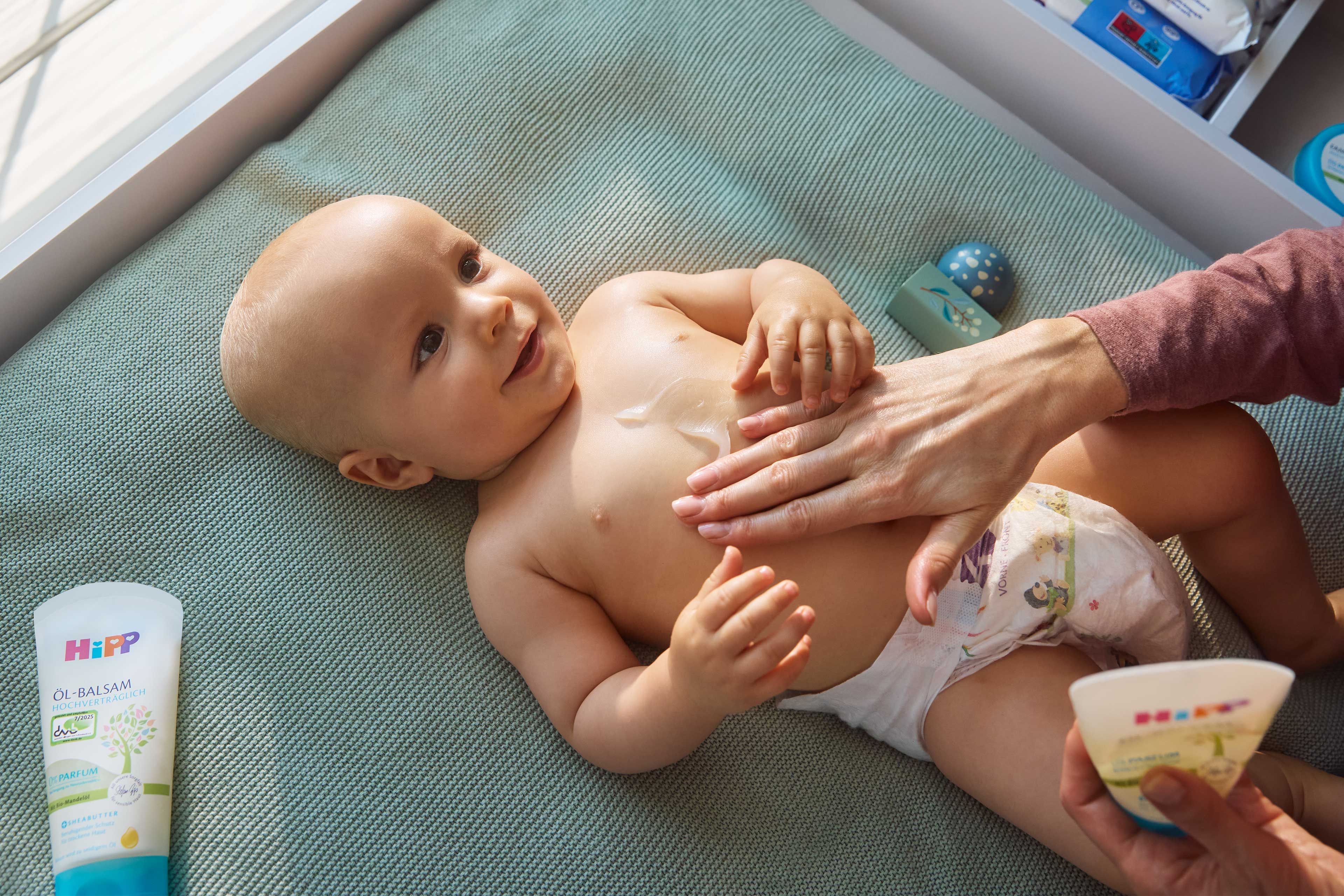 Lächelndes Baby auf der Wickelauflage, wird mit HiPP Babypflege sanft am Bauch eingecremt.