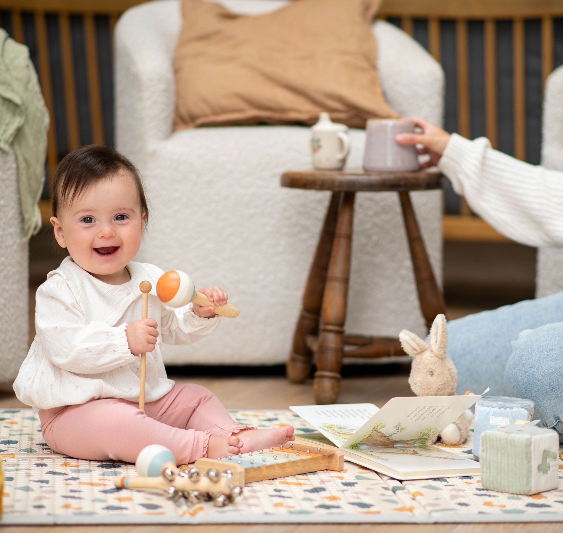 Smiling baby sitting unsupported on a play mat, playing with toys while a parent sits nearby