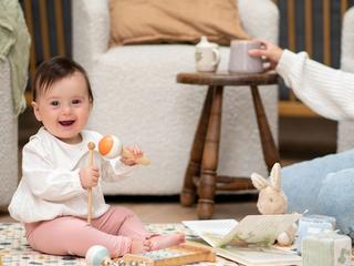 Smiling baby sitting unsupported on a play mat, playing with toys while a parent sits nearby