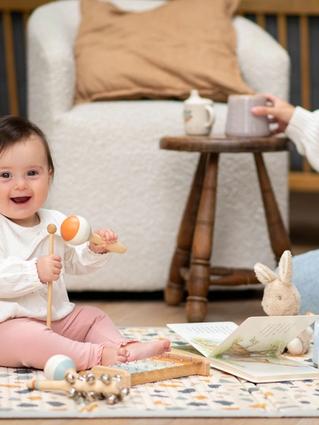 Smiling baby sitting unsupported on a play mat, playing with toys while a parent sits nearby
