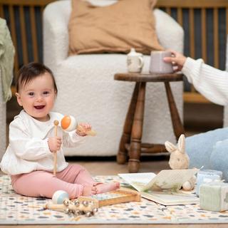 Smiling baby sitting unsupported on a play mat, playing with toys while a parent sits nearby
