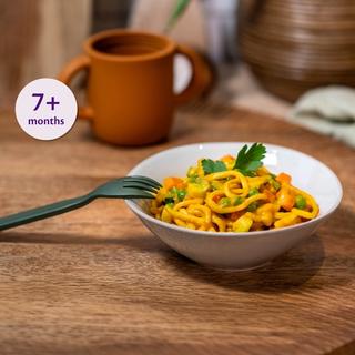 Bowl of veg and chicken noodles with peas and carrots on a wooden table, labelled suitable for babies from 7+ months