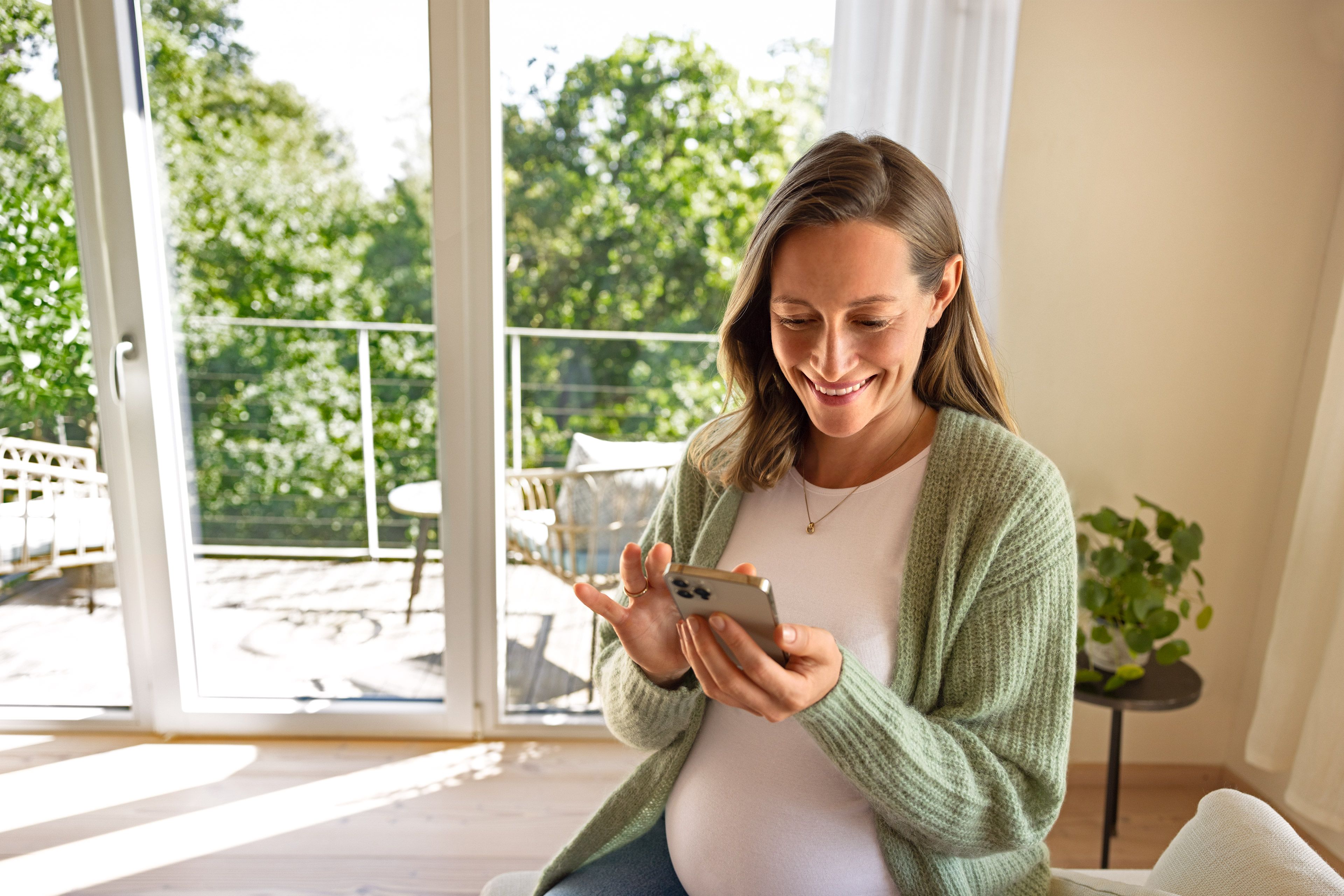 Lächelnde Schwangere nutzt zu Hause ihr Smartphone für hilfreiche Checklisten und Tools.