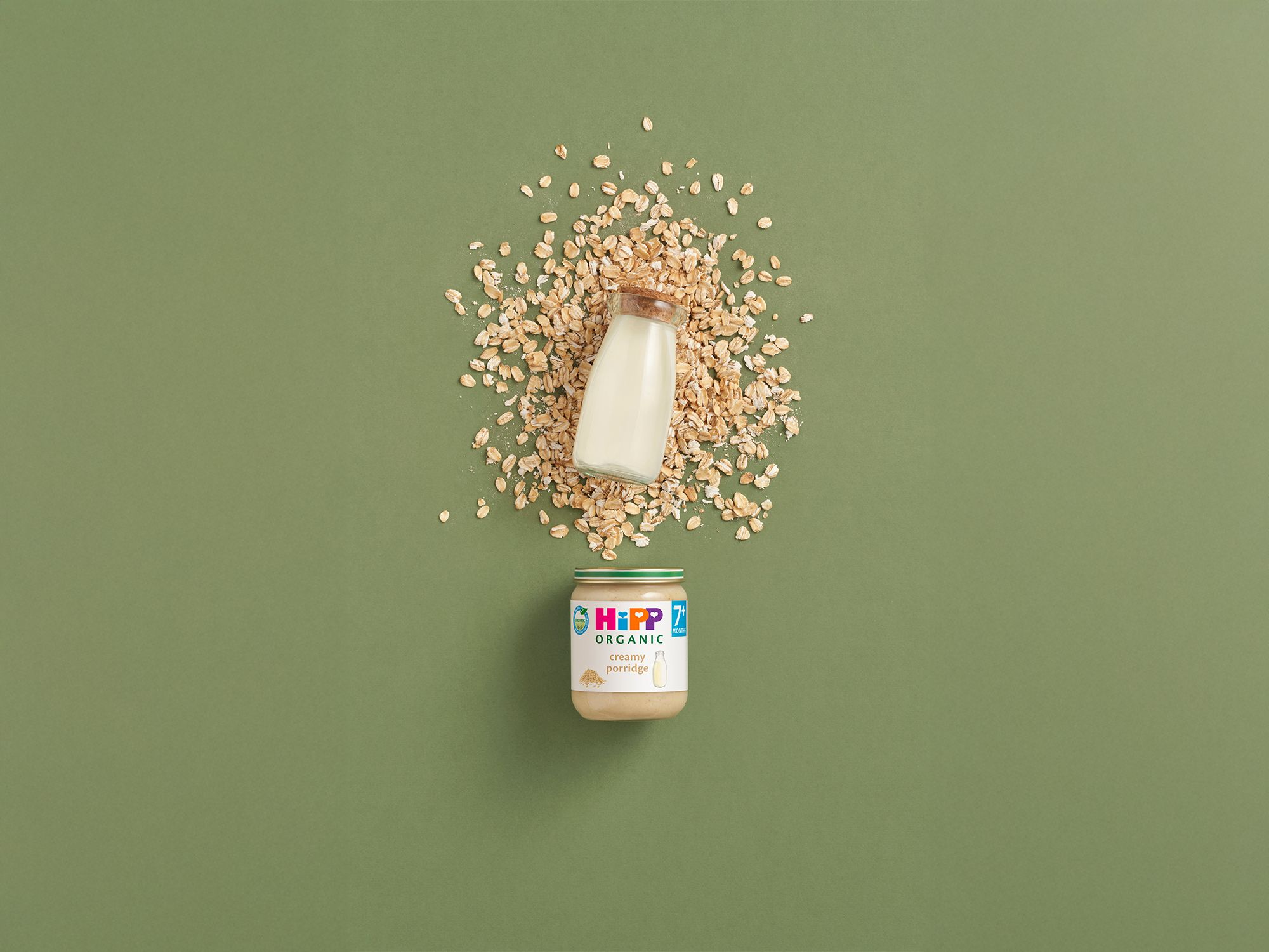 HiPP Organic Creamy Porridge jar with a bottle of milk and scattered oats on a green background