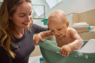 Kleinkind mit Schaum auf dem Körper sitzt in einer Badewanne, daneben eine Frau mit langen Haaren
