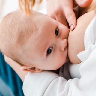 Ein Baby, das von seiner Mutter gestillt wird, wobei das Gesicht des Babys gut sichtbar ist und die Hand der Mutter sanft den Kopf des Babys berührt.