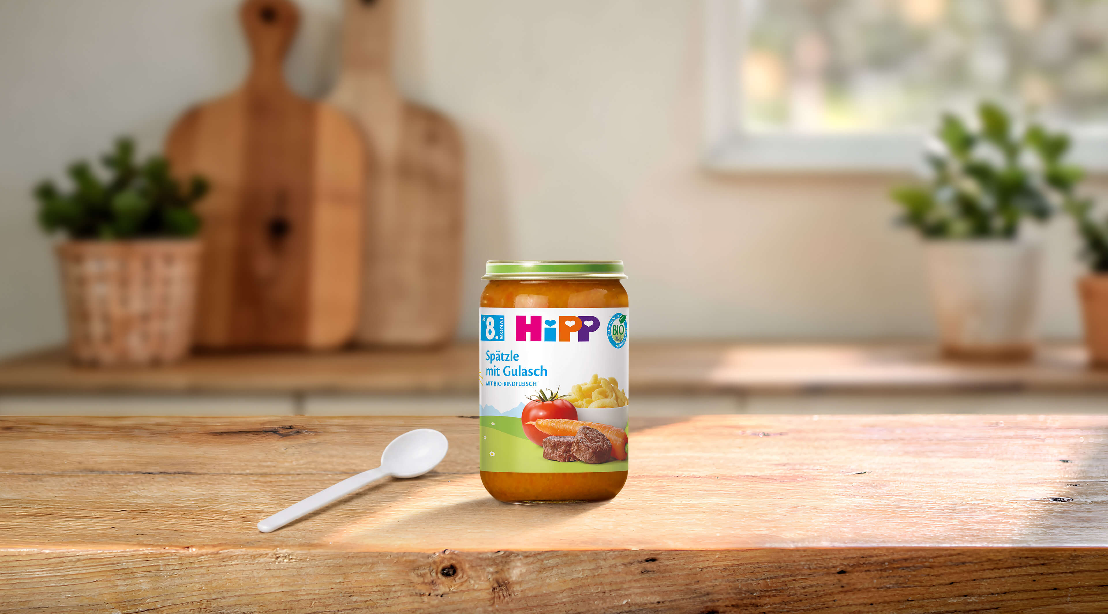 HiPP Bio Babygläschen „Spätzle mit Gulasch“ ab 8. Monat auf Holztisch, daneben ein Löffel in heller, gemütlicher Küche