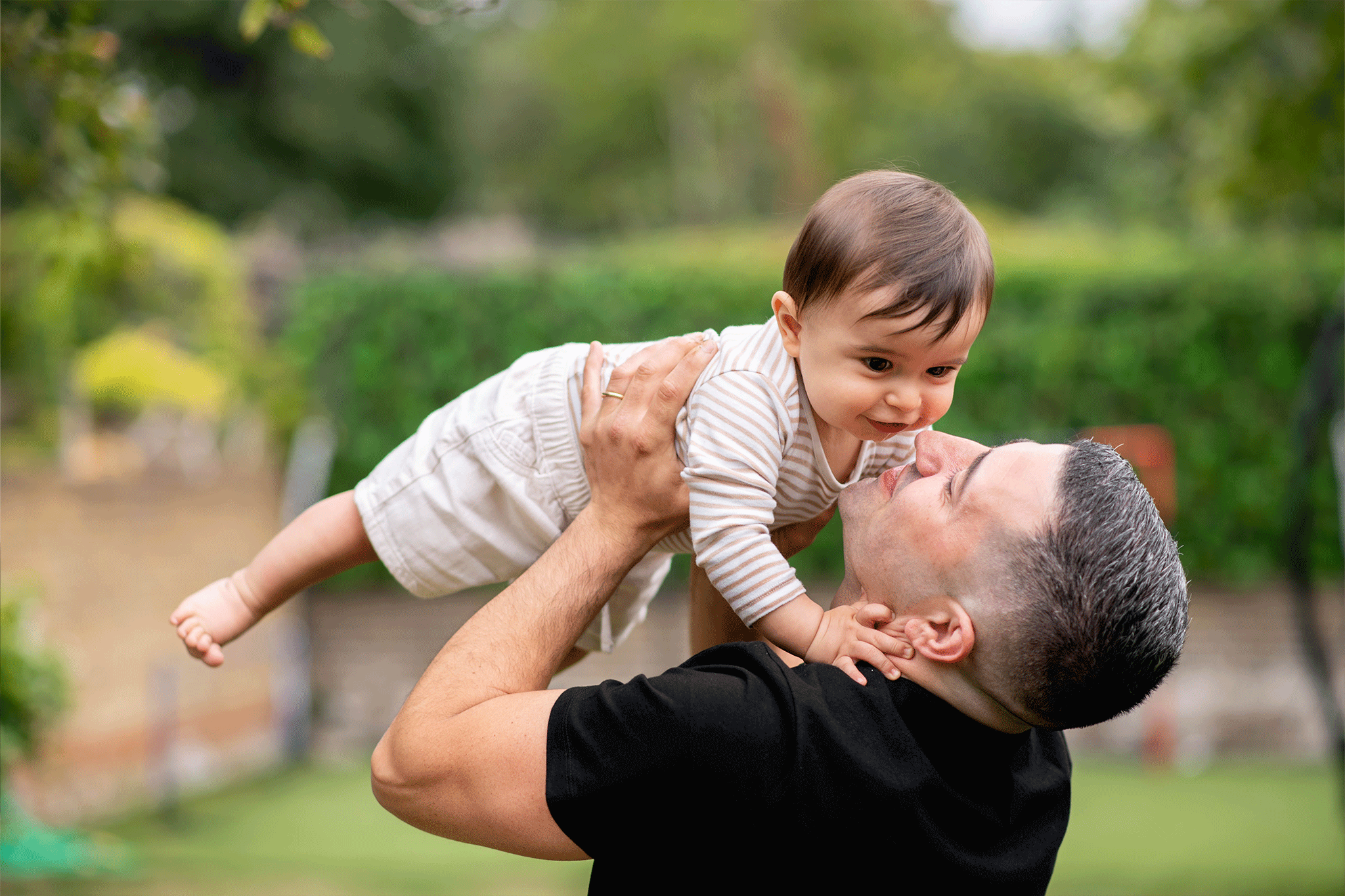 A father in a black t-shirt lifts his smiling baby horizontal in the air outside in a lush, green yard. The baby is wearing a light brown and white striped long-sleeve shirt with beige shorts and looks happily down at their father, who grins back.