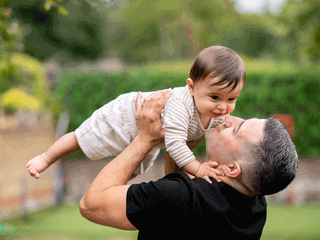 A father in a black t-shirt lifts his smiling baby horizontal in the air outside in a lush, green yard. The baby is wearing a light brown and white striped long-sleeve shirt with beige shorts and looks happily down at their father, who grins back.