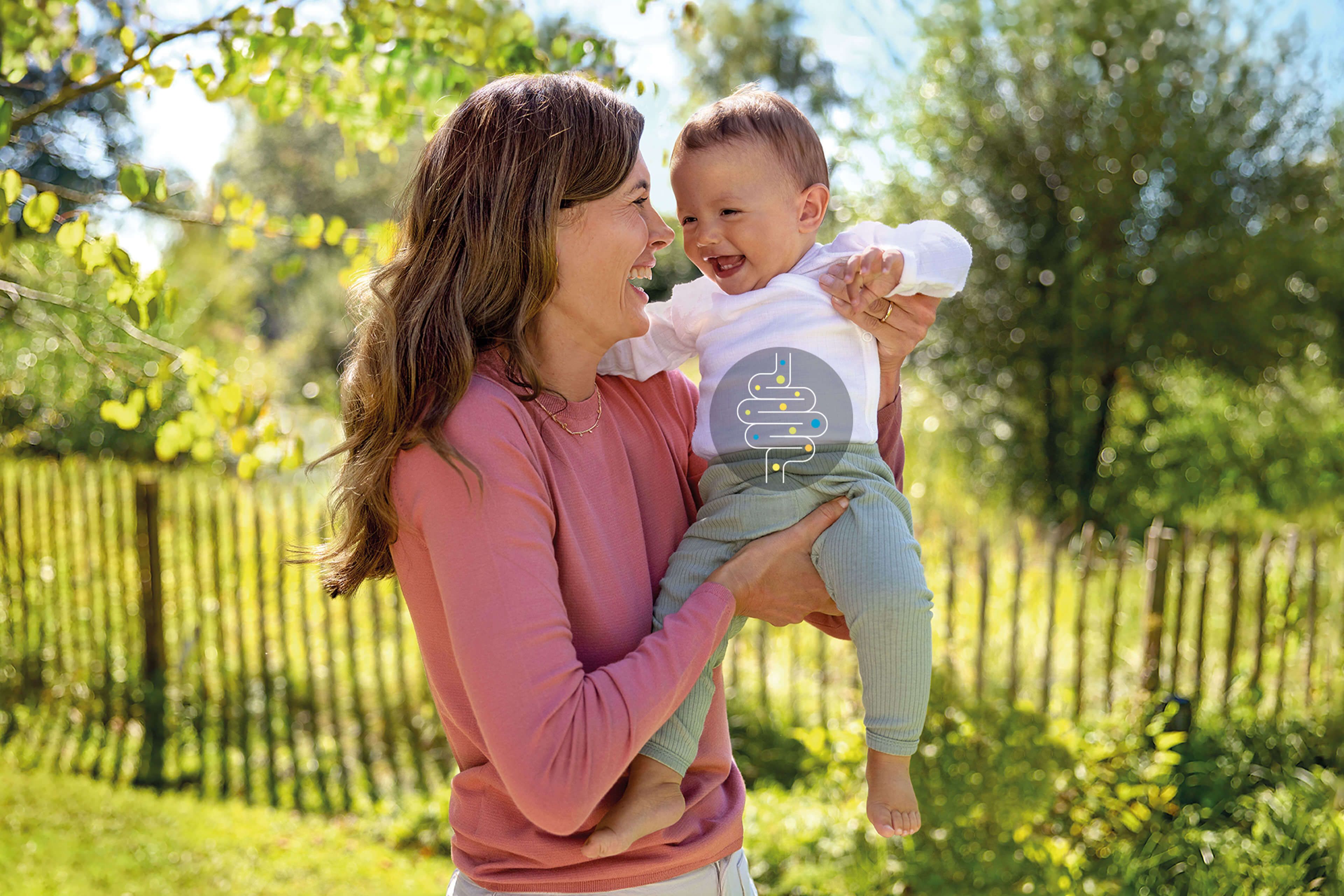 Lachende Mutter hält ihr Baby im grünen Garten, auf dem Body ein Bäuchlein-Symbol und der Text: „Alles, was dein Bäuchlein braucht.“