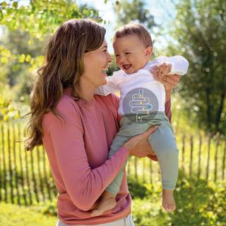 Lachende Mutter hält ihr Baby im grünen Garten, auf dem Body ein Bäuchlein-Symbol und der Text: „Alles, was dein Bäuchlein braucht.“