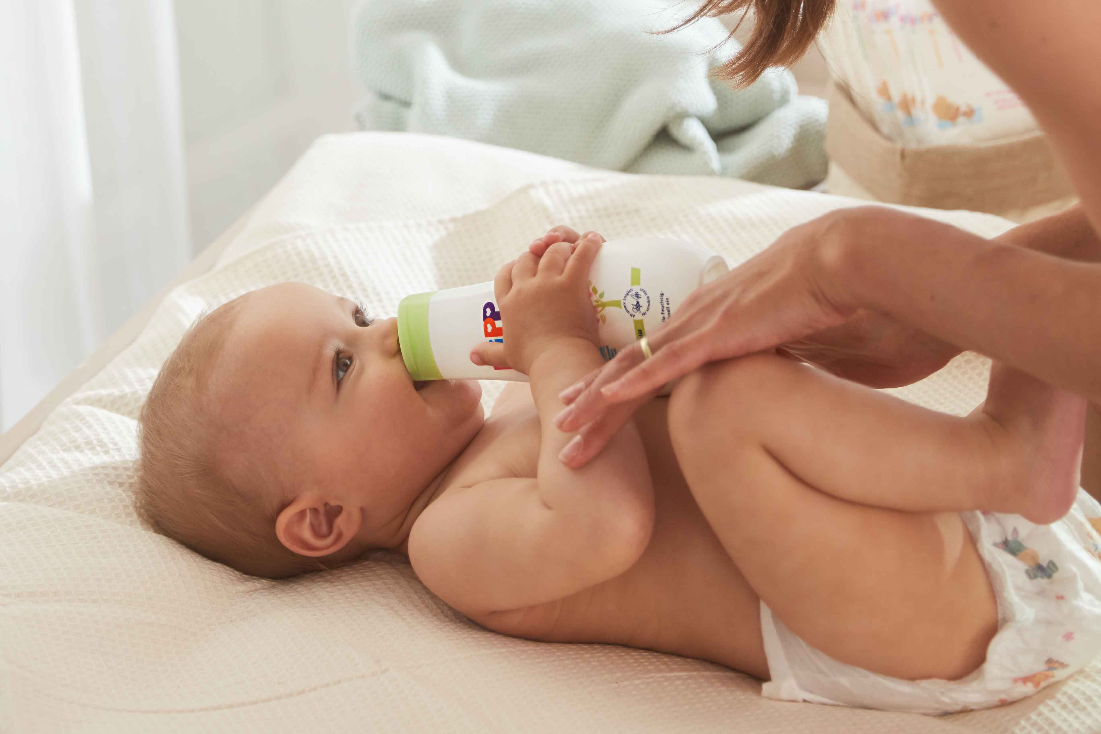 Baby liegt auf Wickeltisch und trinkt aus Flasche während eine Person Hände auf den Bauch legt