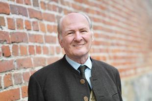 Älterer Mann in traditioneller dunkler Tracht lächelt vor einer Ziegelsteinwand, Porträt zum Unternehmerpreis Pfaffenhofen.