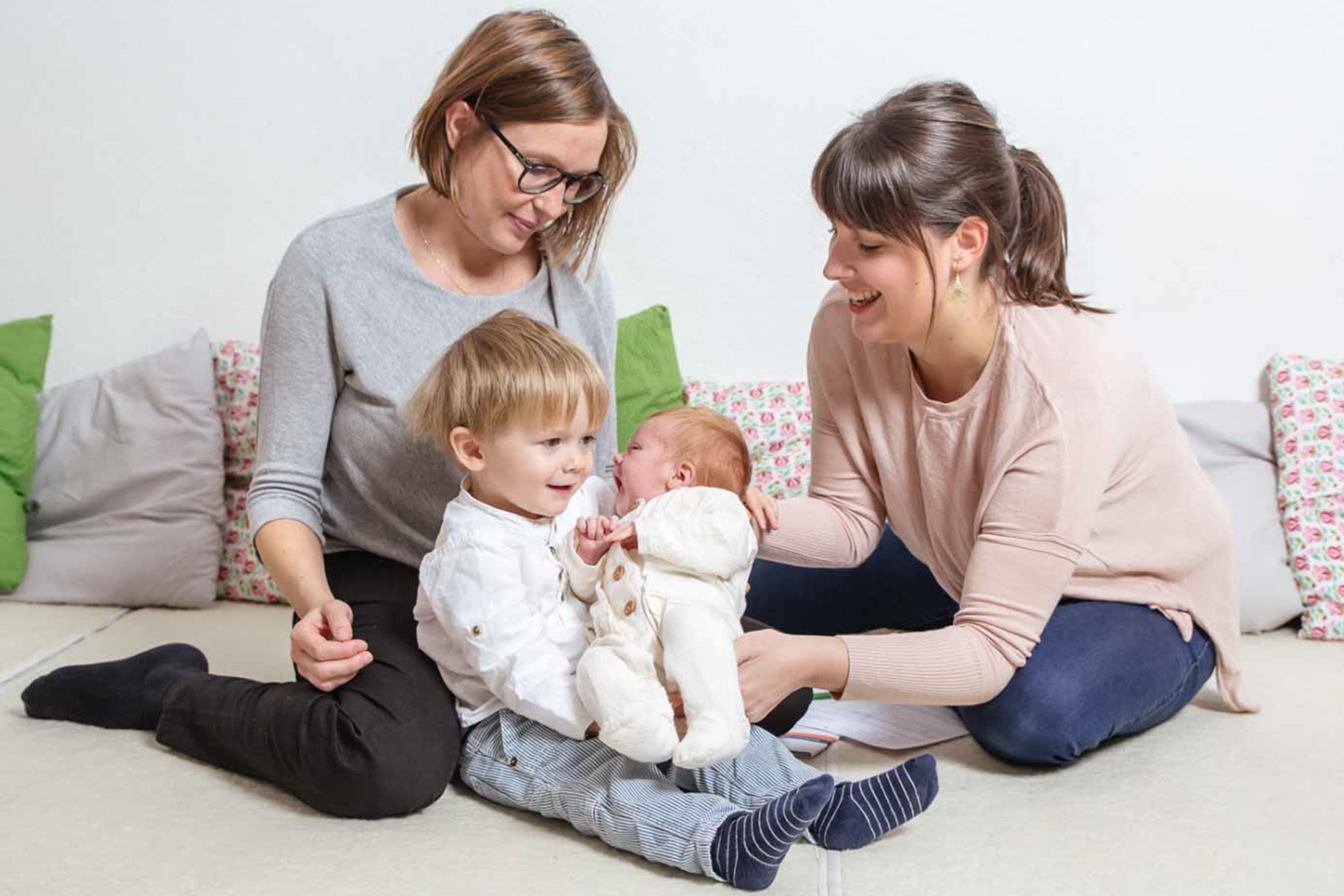 Zwei Frauen, eine mit einem Säugling und die andere mit einem Kleinkind, sitzen auf einem Sofa mit grünen und weißen Kissen.