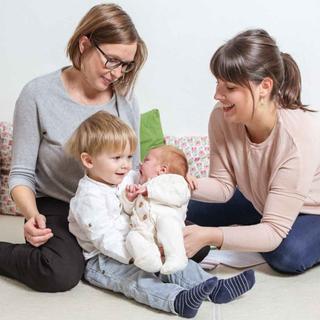 Zwei Frauen, eine mit einem Säugling und die andere mit einem Kleinkind, sitzen auf einem Sofa mit grünen und weißen Kissen.