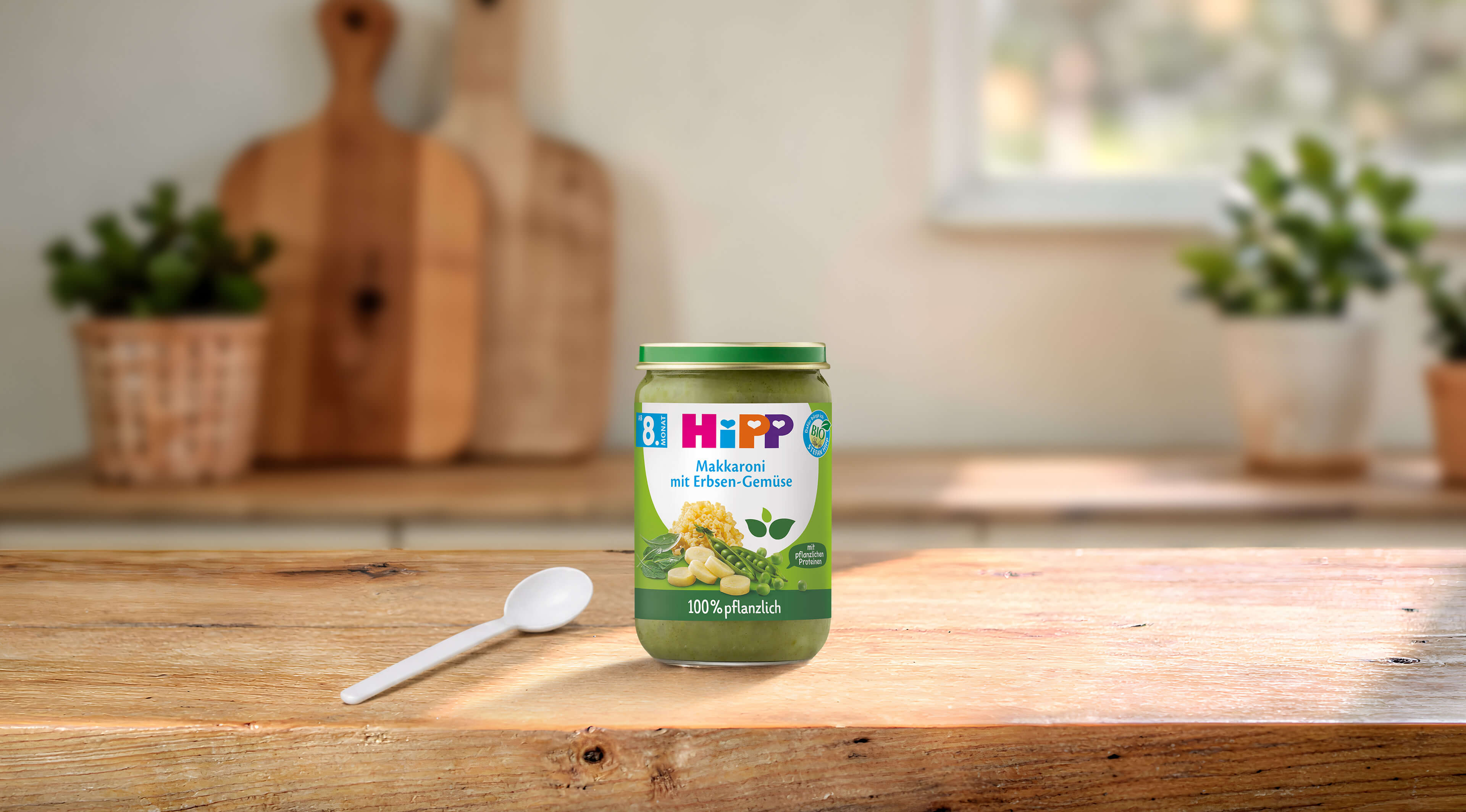 HiPP Bio-Gläschen „Makkaroni mit Erbsen-Gemüse“, 100 % pflanzliche Babykost ab dem 8. Monat auf einem Holztisch mit Löffel in gemütlicher Küche