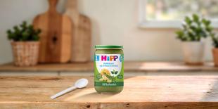 HiPP Bio-Gläschen „Makkaroni mit Erbsen-Gemüse“, 100 % pflanzliche Babykost ab dem 8. Monat auf einem Holztisch mit Löffel in gemütlicher Küche