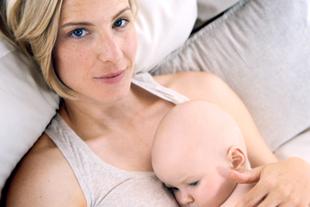 Eine Frau mit blondem Haar und blauen Augen, die ein Baby an sich drückt, das auf einer weichen Unterlage liegt.