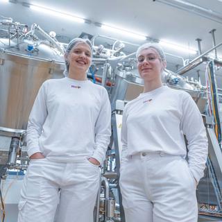 Zwei Auszubildende in weißer Hygienekleidung stehen lächelnd vor modernen Lebensmittel-Produktionsanlagen.