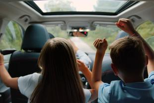 Zwei Kinder, ein Mädchen und ein Junge, sitzen mit erhobenen Händen in einem Auto und scheinen eine Fahrt zu genießen.