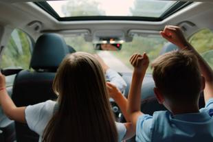 Zwei Kinder, ein Mädchen und ein Junge, sitzen mit erhobenen Händen in einem Auto und scheinen eine Fahrt zu genießen.