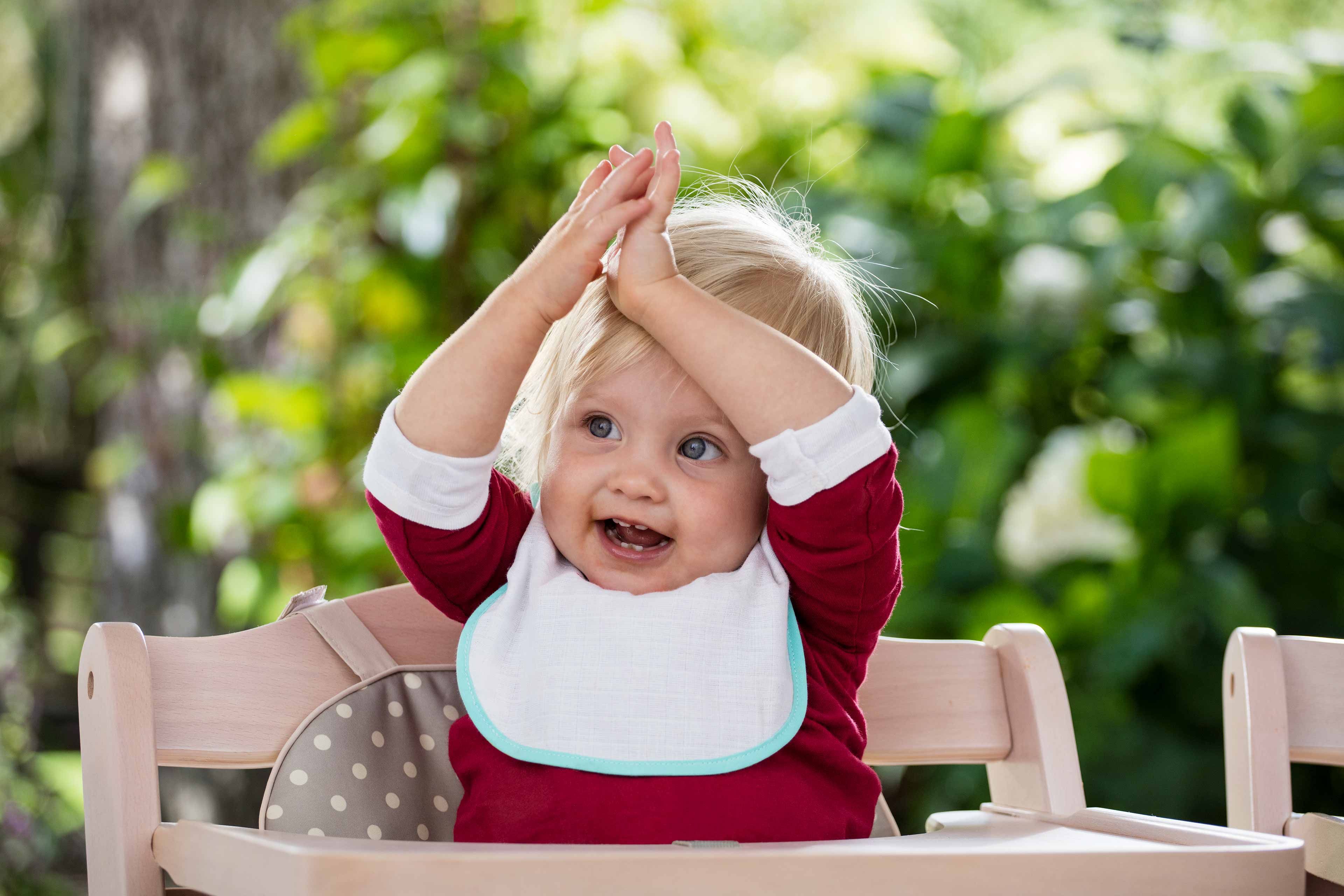 Ein kleines Kind mit blondem Haar, das ein rotes Hemd und ein weißes Lätzchen trägt, sitzt in einem hölzernen Hochstuhl mit einem gemusterten Kissen und hebt beide Hände in einer spielerischen Geste über seinen Kopf.